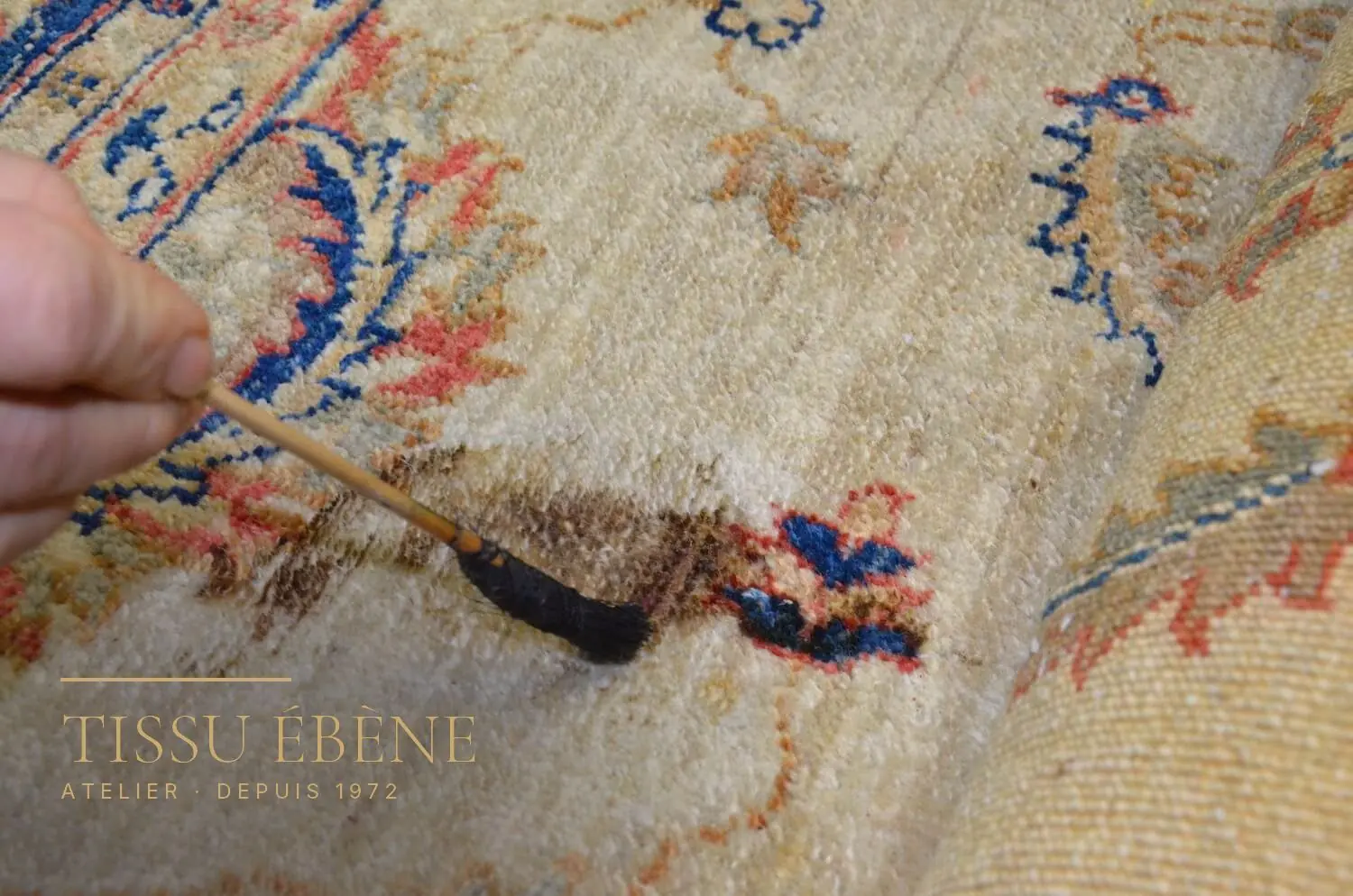 Pinceau posé sur une tache brune d’un tapis à fond ivoire, motifs bleus et corail