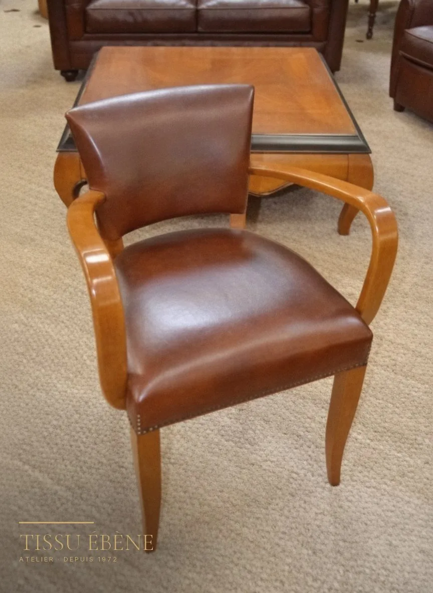 Fauteuil bridge en cuir brun et bois clair, assise cloutée, posé sur une moquette beige