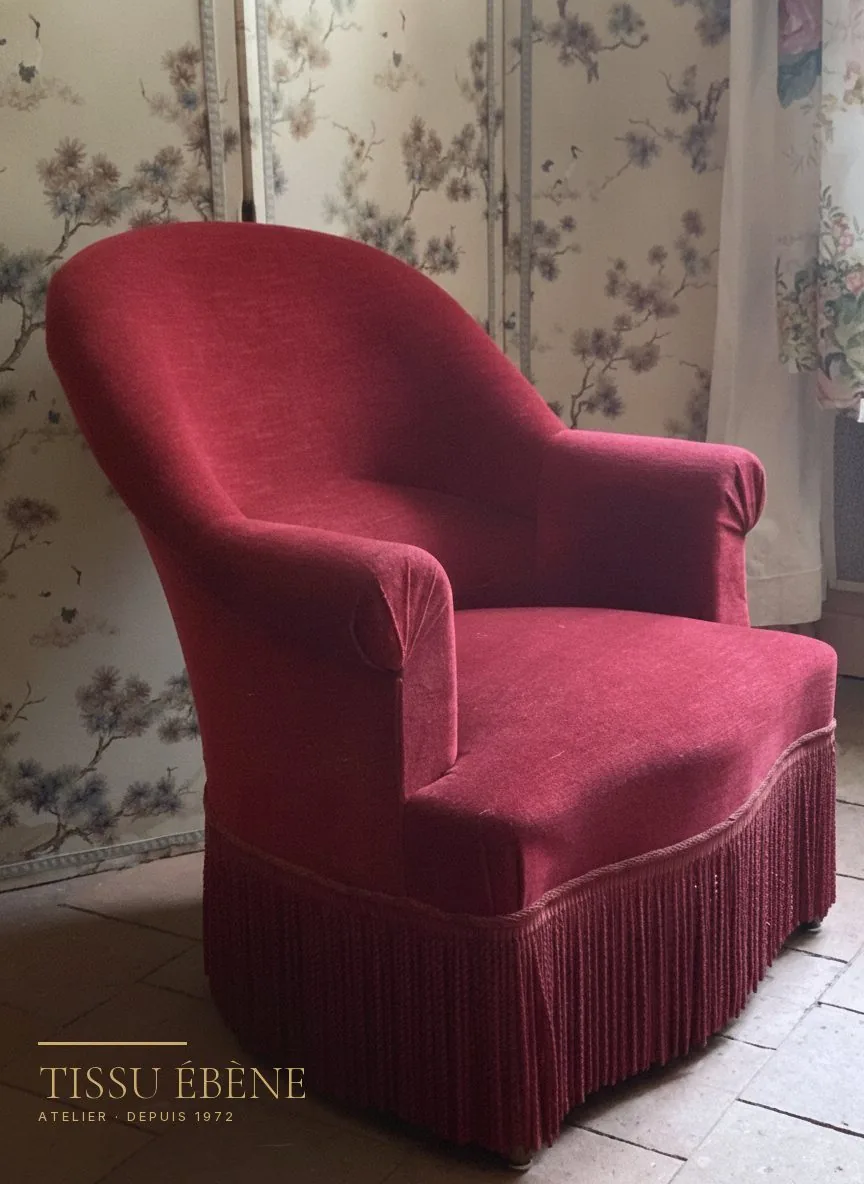 Fauteuil crapaud en velours rouge à franges devant un papier peint fleuri