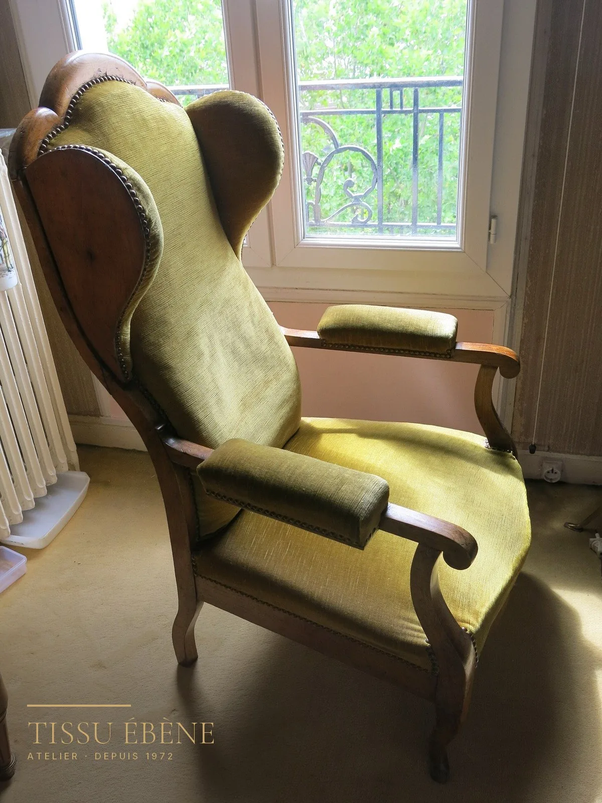 Fauteuil à oreilles ancien garni d’un velours vert olive, bois clair et clous de tapissier, devant une fenêtre